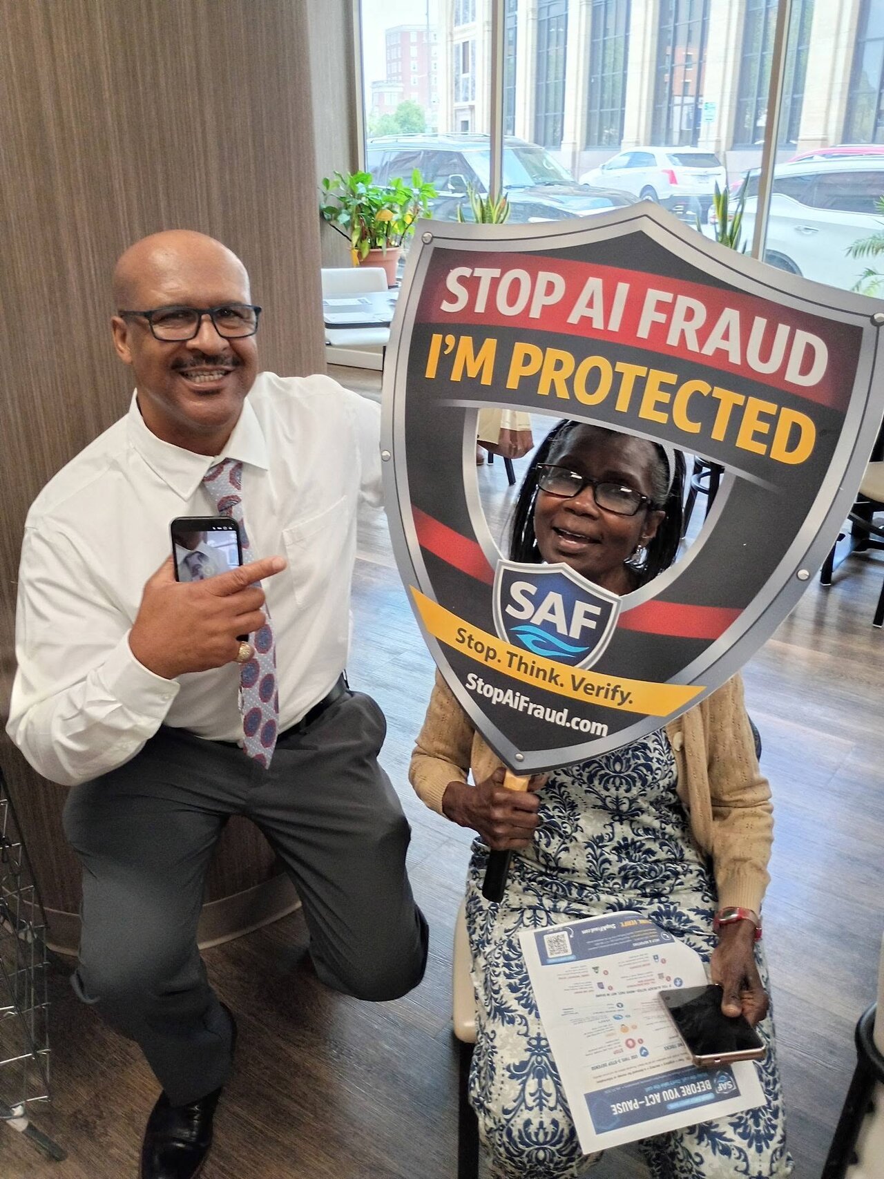 StopAiFraud founder Gary Williams kneels beside a seated attendee holding a "Stop AI Fraud — I’m Protected" shield during the Lifespan Walk Café presentation.