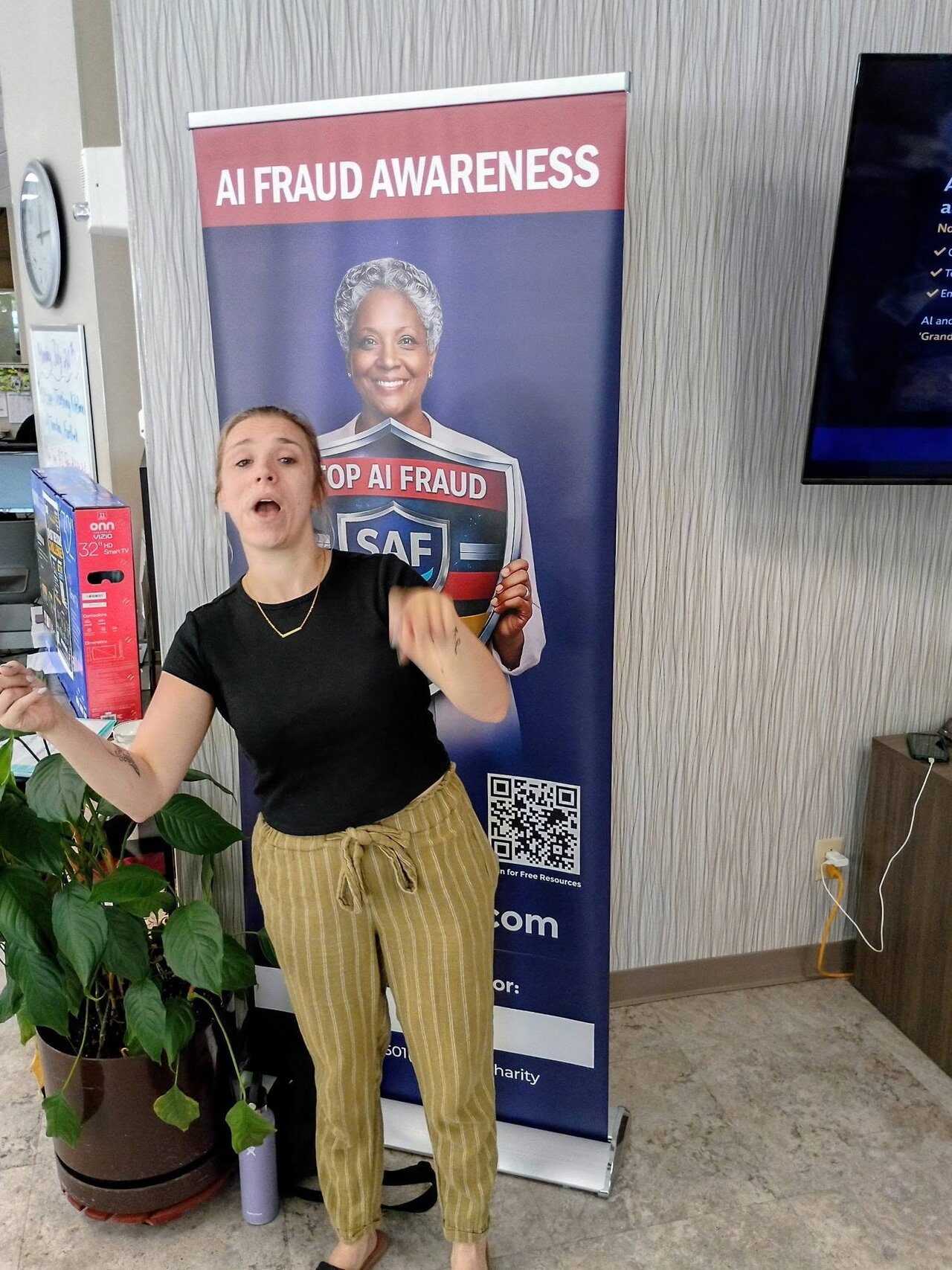 A participant speaks and gestures in front of the AI Fraud Awareness banner during the interactive StopAiFraud presentation at Lifespan Walk Café in Rochester.