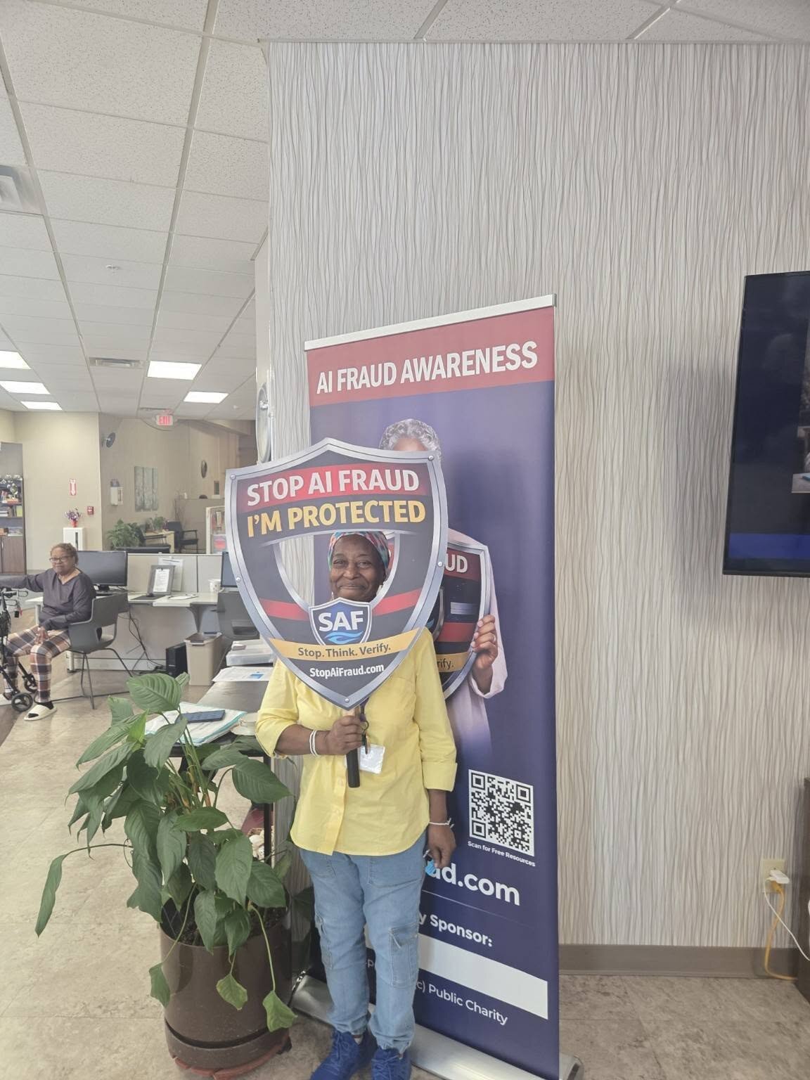 A woman holds up a "Stop AI Fraud — I’m Protected" shield in front of the AI Fraud Awareness banner at the Lifespan Walk Café presentation in Rochester.