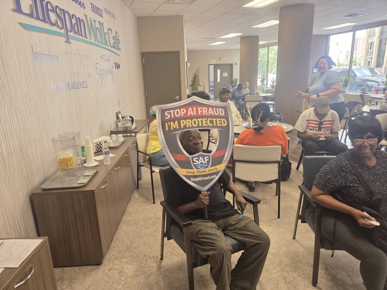 A seated attendee holds a "Stop AI Fraud — I’m Protected" shield during the community AI fraud awareness presentation at Lifespan Walk Café in Rochester.