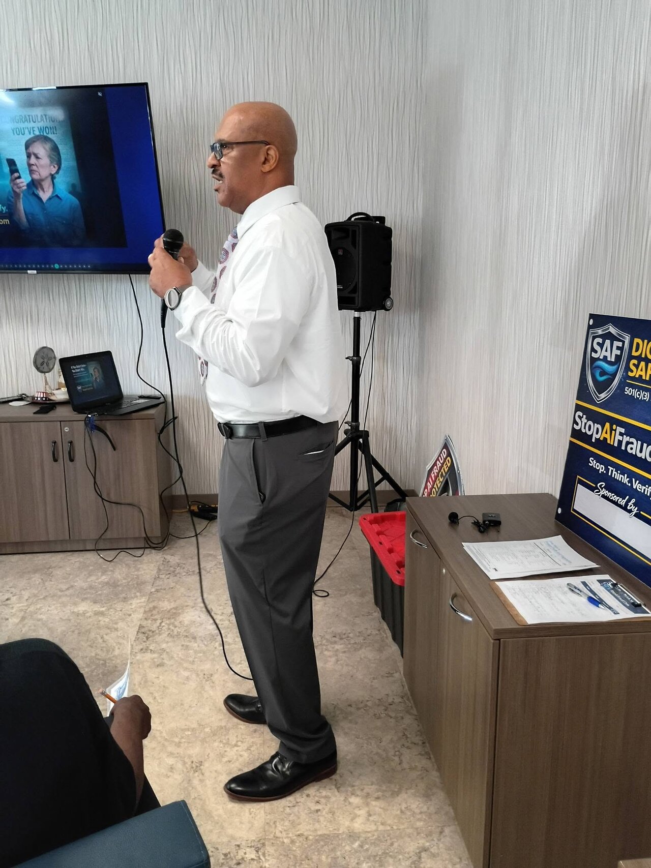StopAiFraud founder Gary Williams speaking with a microphone at Lifespan Walk Café, demonstrating a "Congratulations, You’ve Won" scam example on screen to older adults.